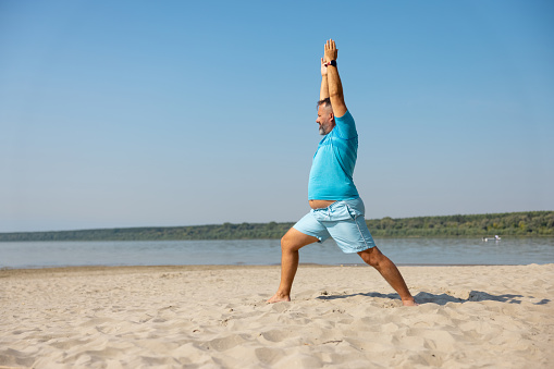 Mid adult man in blue shorts and T-shirt doing yoga exercise on sandy beach, arms raised, hands clasped, lunge posture, High Crescent pose