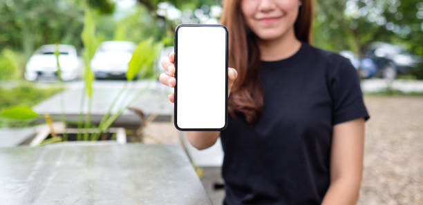 a woman holding and showing a mobile phone with blank white screen in the outdoors stock photo