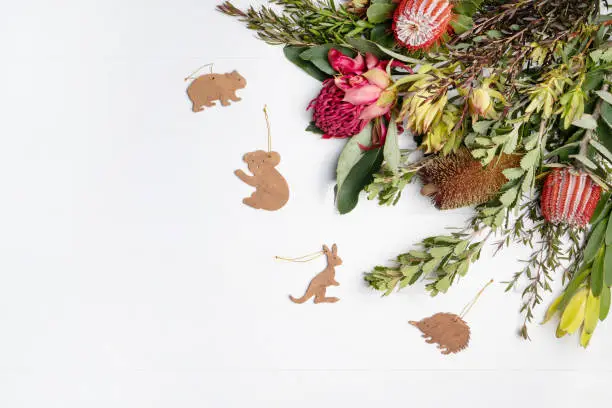 Beautiful flat lay floral arrangement of mostly Australian native flowers, Purple and Red Banksia, Red Waratah and green foliage, decorated by native wooden animals, on a white background. Could be a Christmas background.