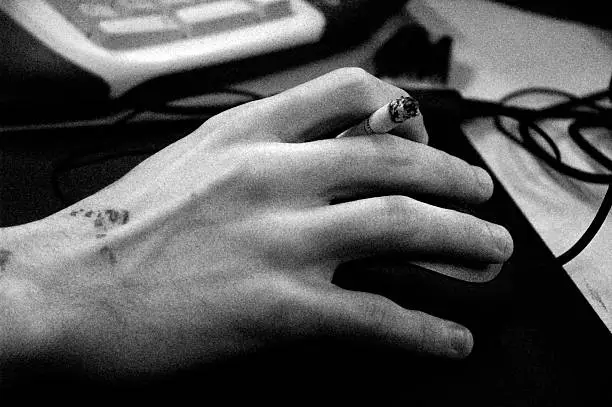 Hand holding a computer mouse and a cigarette, part of keyboard in the background. Grainy Black&White.
