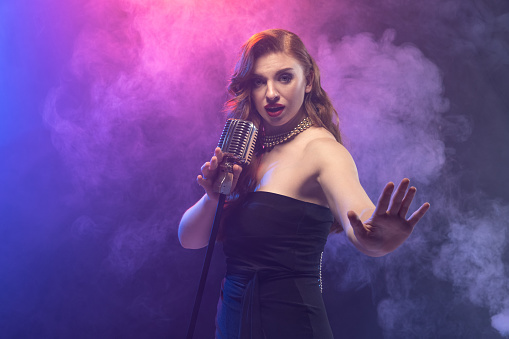 Burlesque singer performing on stage. Woman with retro hairstyle isolated over gradient blue purple background in neon light with stage smoke. Concept of art, music, concert, inspiration, culture, ad