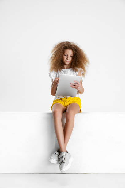 one little cute curly girl in casual clothes sitting on big box with digital tablet, gadget isolated on white studio background. - surprise child little girls shock imagens e fotografias de stock