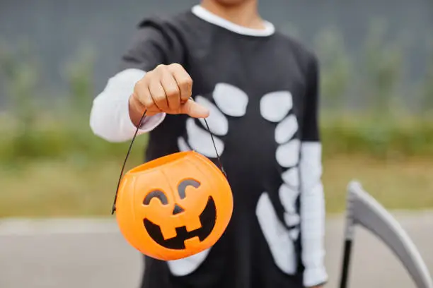Close up of unrecognizable teenage boy holding Halloween pail to camera and wearing costume, trick or treat background