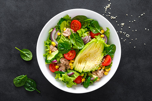 Avocado and tuna fresh vegetable salad with tomato, cucumber corn, onion, lettuce and spinach. Healthy and detox food concept. Ketogenic diet. Buddha bowl dish on black background, top view.