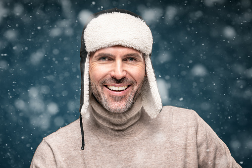 Portrait of handsome man wearing winter cap , posing over snow flakes, looking at camera and smiling. Closeup face of cheerful guy wearing warm clothes.