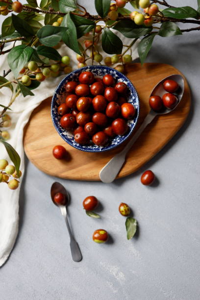 Fresh small jujubes in a rustic ceramic bowl with a blue floral pattern on a light gray rough concrete floor. Around the jujube bowl, there are tree branches with green-yellow berries and green leaves. Hünnap.