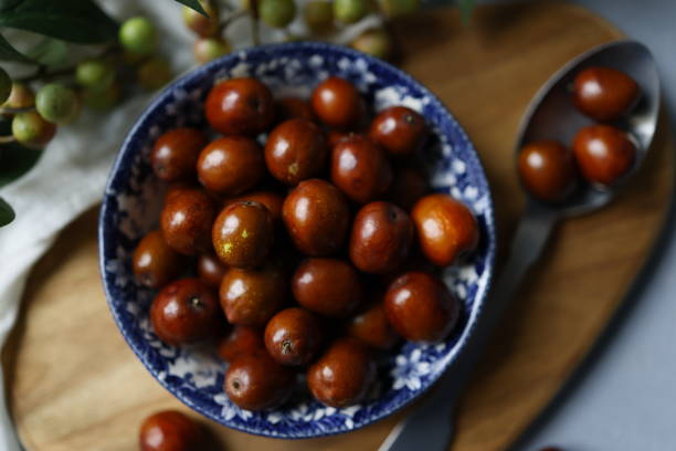 Istanbul, Turkey-September 29, 2021: Small jujubes in a rustic ceramic bowl with a blue floral pattern on a light gray rough concrete floor. Around the jujube bowl, there are tree branches with green-yellow berries and green leaves. The jujube bowl stands on the wooden cutting board. There are little jujubes scattered around. The Turkish name of this fruit is “hünnap”. Shot with Canon EOS R5.