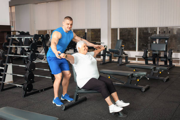 istock pensioner performs an exercise with dumbbells on simulator in gym. coach shows and helps. Sporty healthy lifestyle, senior concept, indoor. Rehabilitation, treatment, improvement of self-awareness. 1343869041