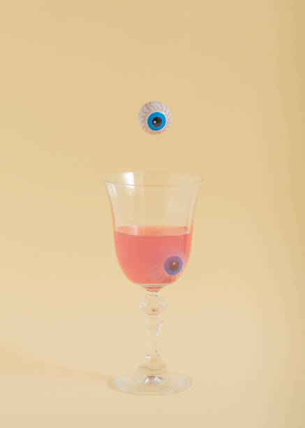 Spooky Halloween concept with an eye falling into a drinking glass. Vertical aesthetic composition, creative season concept. Pastel beige background.