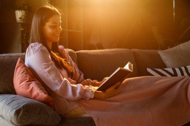 Young woman relaxing on the sofa under a cozy blanket, reading a book