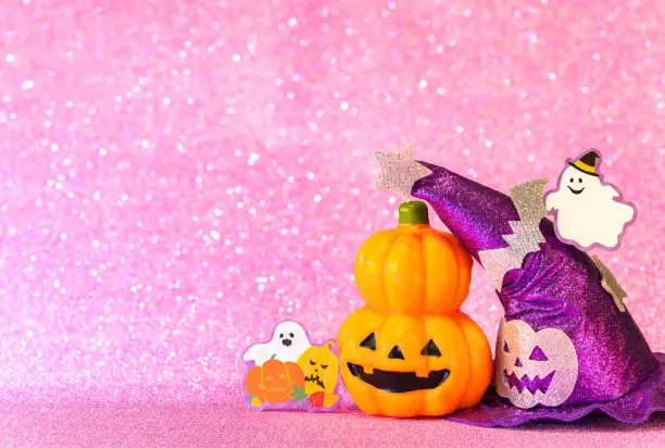 Photo of Halloween jack o lantern pumpkin and witches hat with smiling ghost  on  a glittering pink background.