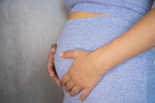 A pregnant woman is standing in a blue suit and holding her tummy with her hands on a gray background. High quality photo