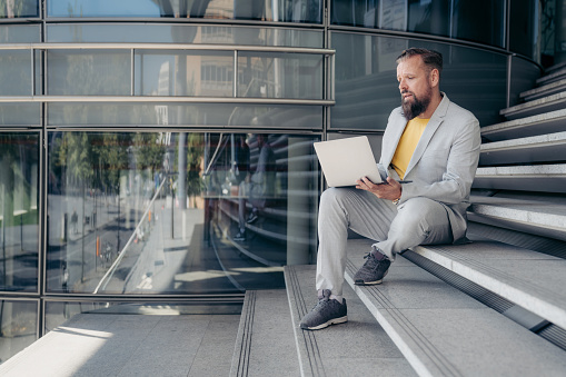 Mature businessman sitting on stairs in front of his office building and working on laptop. Executive using laptop computer sitting on steps of office building.
