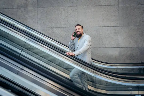 Businessman standing moving up on escalator while talking on phone in city. Happy businessman standing on stairs and using smart phone.