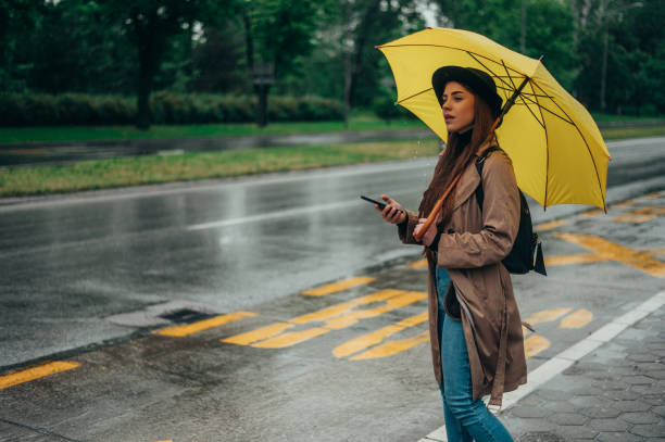 istock Woman holding yellow umbrella and using smartphone while on a bus stop 1343926142