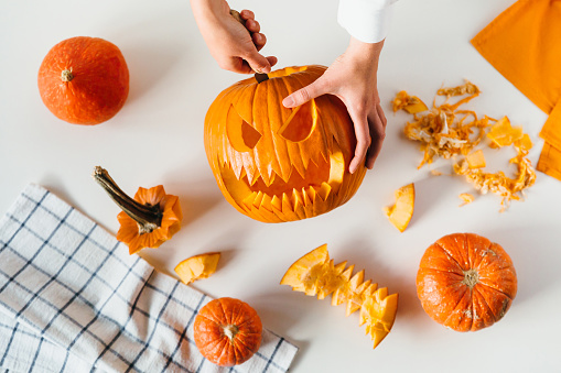 Happy halloween, decoration and holidays concept. Young woman hands with knife carving pumpkin or jack-o-lantern on white table background at home. Close-up, top view, flat lay.