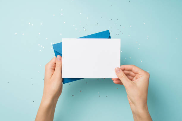 foto in prima persona con vista dall'alto di mani che tengono una busta blu e una carta bianca su paillettes su uno sfondo blu pastello isolato con spazio vuoto - cartolina di auguri foto e immagini stock