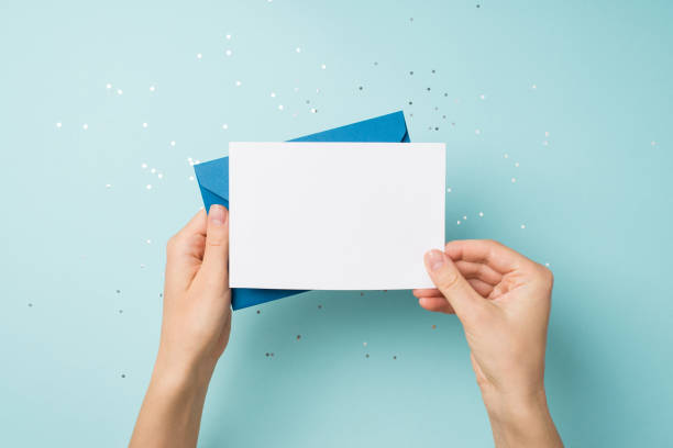 空白のスペースを持つ孤立したパステルブルーの背景にスパンコールの上に青い封筒と白いカードを保持している手の一人称トップビュー写真 - グリーティングカード ストックフォトと画像