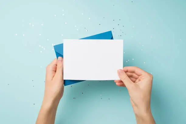 Photo of First person top view photo of hands holding blue envelope and white card over sequins on isolated pastel blue background with blank space