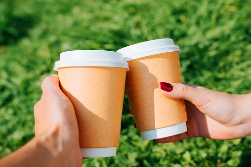 Close-up of hand holding two disposable cups of coffee, woman's and man's hand clink glasses outdoors on green lawn. Selective focus on right cup.