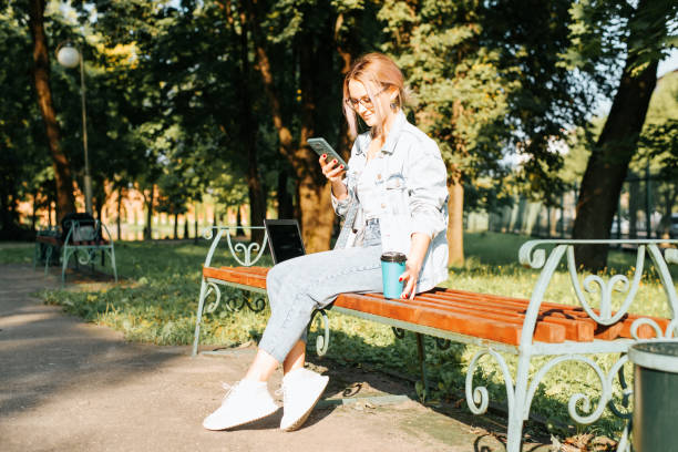 istock Smiling young woman wearing glasses and casual clothes using smartphone while holding disposable cup of coffee and relaxing on park bench. Girl chatting, browsing Internet on mobile phone, outdoors 1343987831