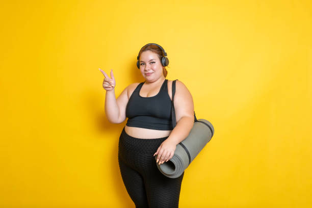 overweight and fitness young caucasian hispanic model in sportswear with headphones showing yoga mat pointing to the side - athlete muscular build yoga female imagens e fotografias de stock