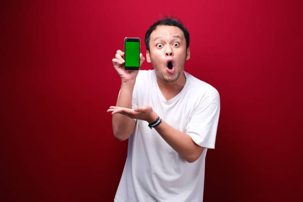 istock Shock Young Asian man pointing on green blank space on smartphone on red background. 1344008479