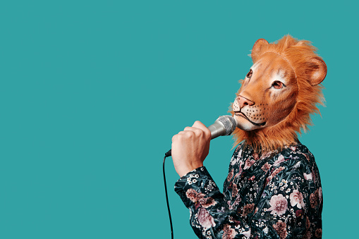 a young man wearing a lion mask speaks into a microphone, against a blue background with some blank space on the left