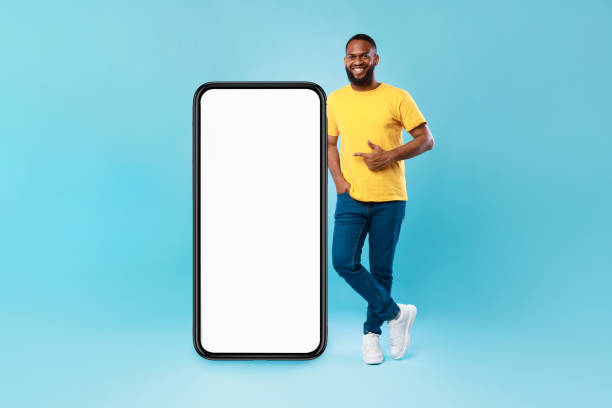 istock Positive Afro guy pointing at big smartphone with mockup for mobile app, recommending cool website over blue background 1344020352