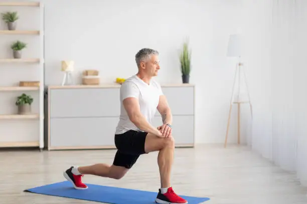 Home fitness. Motivated mature man doing lunges in living room interior, free space, banner design. Man working out, training leg muscles during coronavirus, staying at home