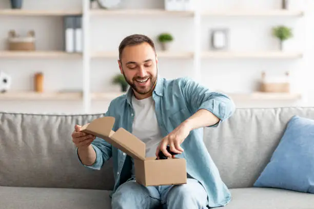 Satisfied buyer. Portrait of happy Caucasian man receiving package, unboxing parcel, taking out gaming joystick, sitting on sofa in living room. Millennial guy feeling happy with online purchase
