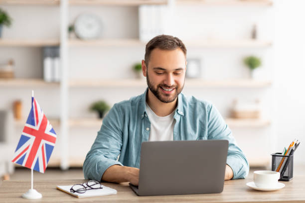 Happy young guy with flag of Great Britain working at desk with laptop in home office Happy young guy with flag of Great Britain working at desk with laptop in home office. Cheerful male student studying British English online for abroad job, education or emigration england stock pictures, royalty-free photos & images