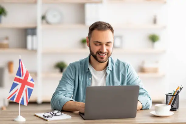 Photo of Happy young guy with flag of Great Britain working at desk with laptop in home office