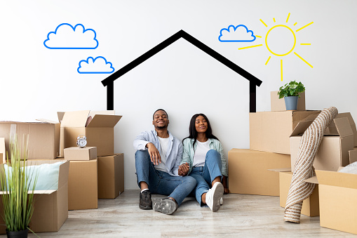 Loving african american spouses relocating to their own home, dreaming about renovation while sitting near white wall with drawn house among cardboard boxes