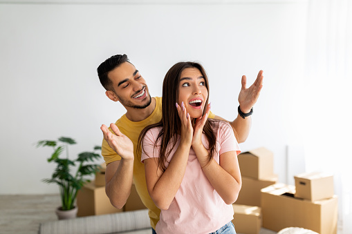 Excited Arab guy surprising his Caucasian girlfriend, moving to their own home together, standing in apartment among carton boxes. Happy diverse couple on first day in their new house