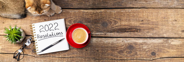 istock New year Resolutions 2022 on desk. 2022 Resolutions with notebook, cute cat, coffee cup, eyeglasses, succulent on wooden background. Goals, planning, strategy, cozy home concept 1344021720