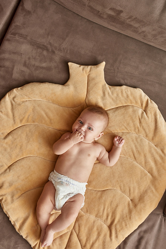 Joyful infant baby on cotton play mat in leaf form, top view. Naked baby sucking fingers while lying alone on floor. Copy space. Handmade leaf play mat from natural linen.