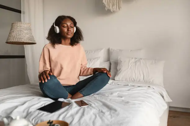 Online Meditation. Peaceful Black Woman Meditating With Eyes Closed Wearing Headphones Sitting In Lotus Position, Listening To Yoga Instructor Via Digital Tablet Relaxing In Bedroom At Home