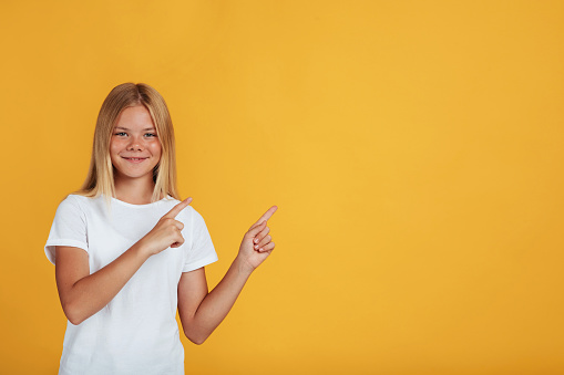 Smiling teen pretty blonde girl pupil in white t-shirt pointing her fingers at empty space, isolated on yellow background. Great advertising, offer and advice, student positive emotions, studio shot