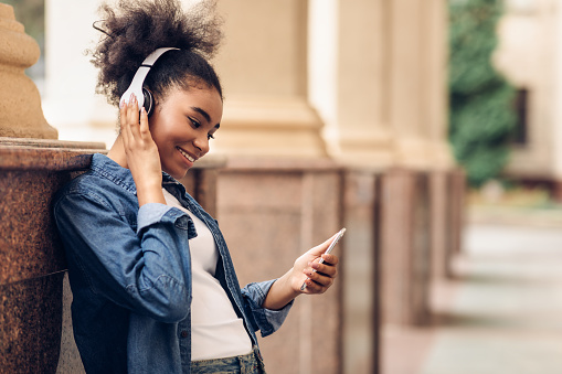 Happy African Teen Girl Using Phone Listening To Music Online Wearing Wireless Headphones Spending Time At University Standing Outdoors. People And Gadgets, Studentship Lifestyle. Side View