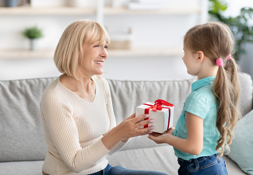 Celebrating Concept. Portrait of cute little girl making surprise for her grandma holding and giving present box, greeting woman with holiday. Granddaughter congratulating smiling mature lady