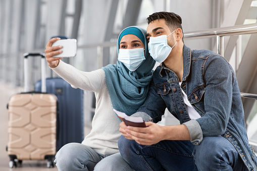 Young muslim couple wearing medical masks taking selfie on smartphone while waiting for flight in airport terminal, happy islamic spouses travelling together during coronavirus pandemic, closeup shot