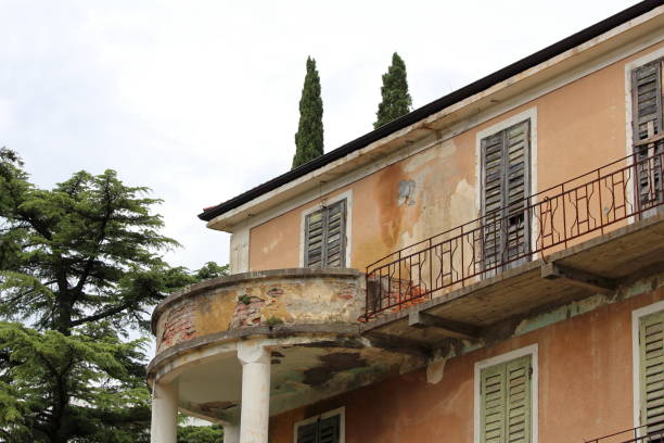 Rounded concrete balcony supported by two white columns next to elongated balcony with rusted metal fence on side of old dilapidated abandoned building with cracked facade and destroyed wooden window blinds surrounded with tall pine trees on cold spring day