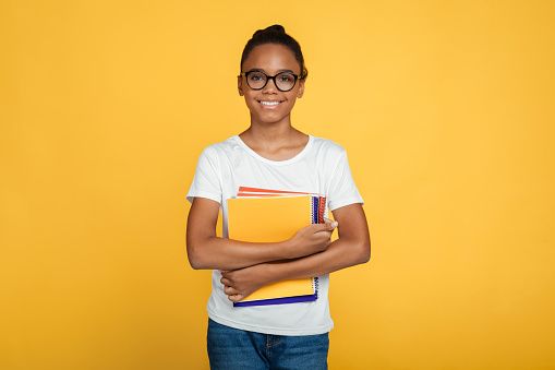 Cheerful teenage african american girl pupil in glasses and casual holds books and ready to learn, isolated on yellow background, free space. Modern education, knowledge, back to school in autumn