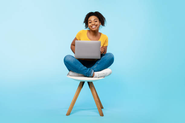 Cheery young black woman working online, sitting on chair and using laptop on blue studio background, full length Cheery young black woman working online, sitting on chair and using laptop on blue studio background, full length. Cheerful African American lady surfing internet on portable pc office chair stock pictures, royalty-free photos & images