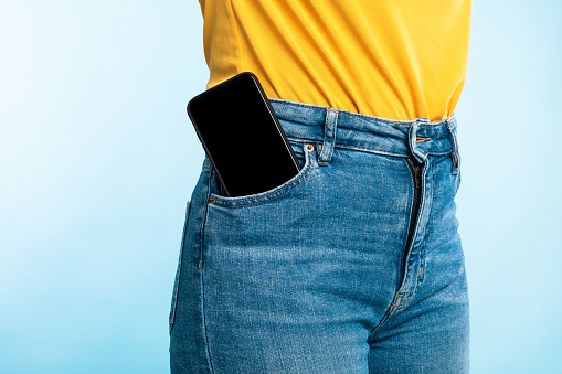 Unrecognizable woman having smartphone with empty screen in her jeans pocket on blue background, mockup