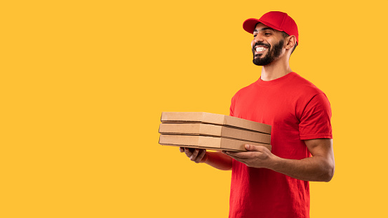 Pizza Delivery. Arabic Courier Man Holding Pizza Boxes Smiling Looking Aside Delivering Food From Pizzeria Posing Standing Over Yellow Background. Panorama, Studio Shot