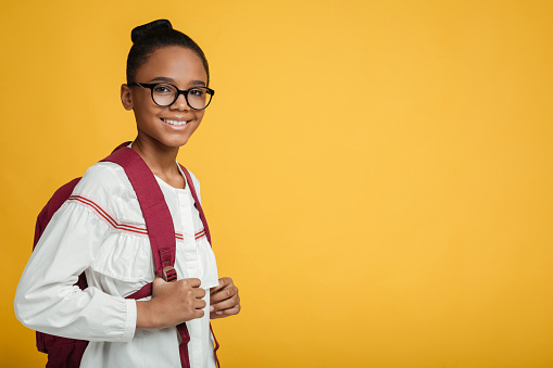 Happy smart teen afro american girl pupil in glasses with backpack look at camera, isolated on yellow background, free space. Modern schoolgirl, back to school after covid-19, education and knowledge