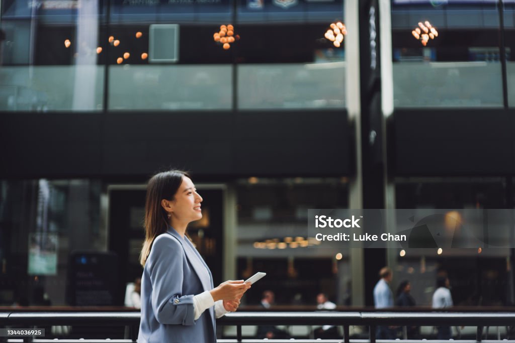 Business on the go Beautiful Asian businesswoman walking in a financial area, she is looking up to the sky and holding a smartphone. Banking Stock Photo