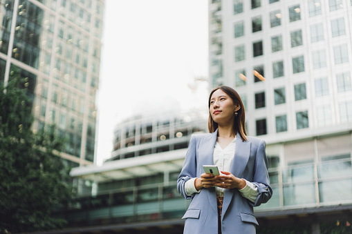 Beautiful Asian businesswoman walking in a financial area, she is looking up to the sky and holding a smartphone.
